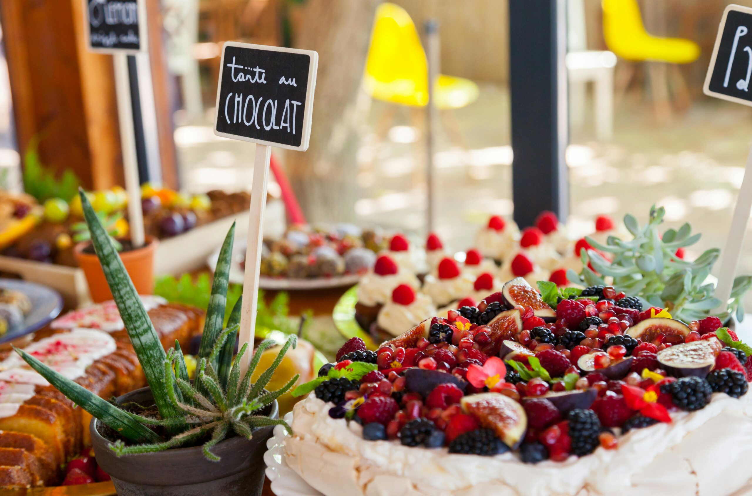 A colorful fruit cake surrounded by an array of desserts on an outdoor table setting.